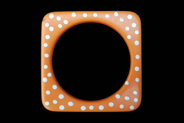 Horizontal top view of 1980s vintage orange and white square sliced polka dot, striped, and translucent solid three (3) bangle bracelet set. Showing the orange with white polka dots bangle alone. It has a small dark mark on the front corner.