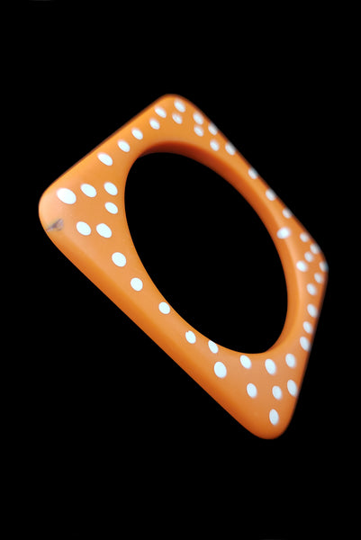 Vertical perspective view of 1980s vintage orange and white square sliced polka dot, striped, and translucent solid three (3) bangle bracelet set. Showing the orange with white polka dots bangle alone. It has a small dark mark on the front corner.