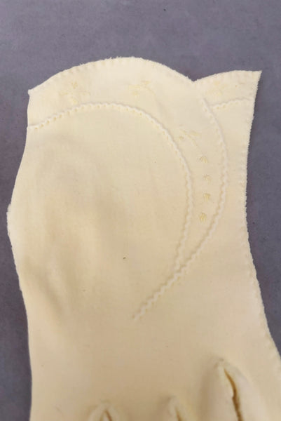 Detail view of 1950s vintage pastel yellow cotton wrist length curved cuff embroidered gloves. Approximately size 7.5. Showing a close-up of the curved cuff with puckered decorative stitching and embroidered sprigs. 