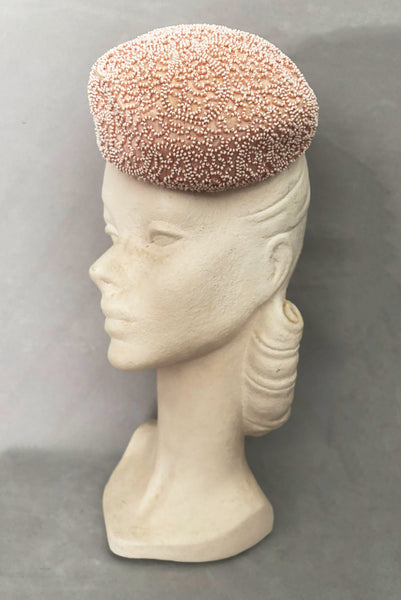 Vertical 3/4 view of 1960s vintage blush pink faille fabric pillbox hat covered in matching glass seed beads by Neiman Marcus.