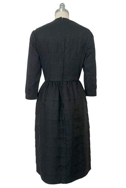 Back view of 1960s vintage crew neck, 3/4 sleeve, bow trimmed black faille cocktail dress, with tiered skirt, size small to medium.