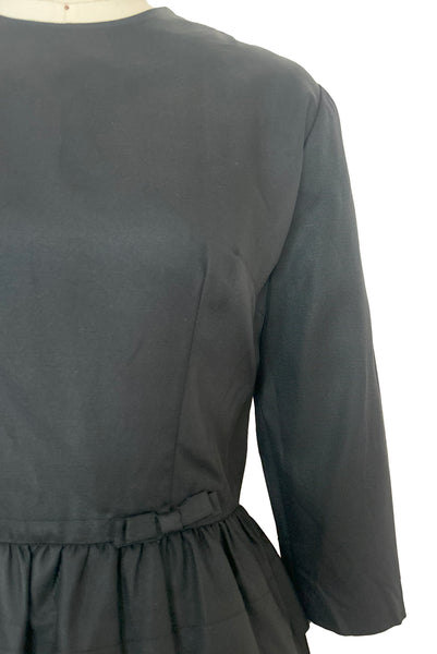 Detail view of 1960s vintage crew neck, 3/4 sleeve, bow trimmed black faille cocktail dress, with tiered skirt, size small to medium. Showing a close up of the bows at the waist.