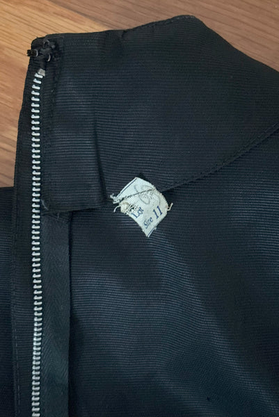 Interior detail view of 1960s vintage crew neck, 3/4 sleeve, bow trimmed black faille cocktail dress, with tiered skirt, size small to medium. Showing a close-up of the size 11 tag.