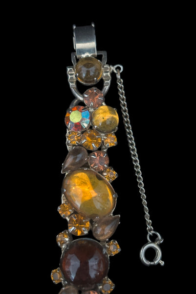 Detail view of 1950s vintage Weiss autumnal orange, brown, and iridescent rhinestone and foil backed glass cabochon gold tone bracelet. Showing some dark spots in the glass cabochons, the hinged clasp, and safety chain.
