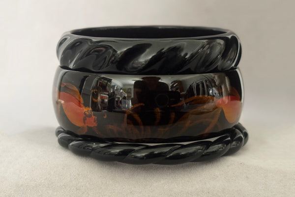 Horizontal side view of 1980s vintage black plastic twisted, ridged, and orange goldfish painted bangle bracelet set of three (3).