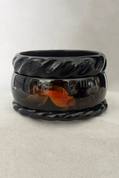 Vertical front view of 1980s vintage black plastic twisted, ridged, and orange goldfish painted bangle bracelet set of three (3).