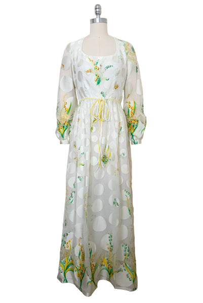 Front view of 1960s vintage white chiffon, satin polka dot, yellow and green lily of the valley print floral maxi dress by Richilene New York for Neiman Marcus, size small to medium. With scoop neck, long full sleeves, elevated waist, and floor length gathered skirt.