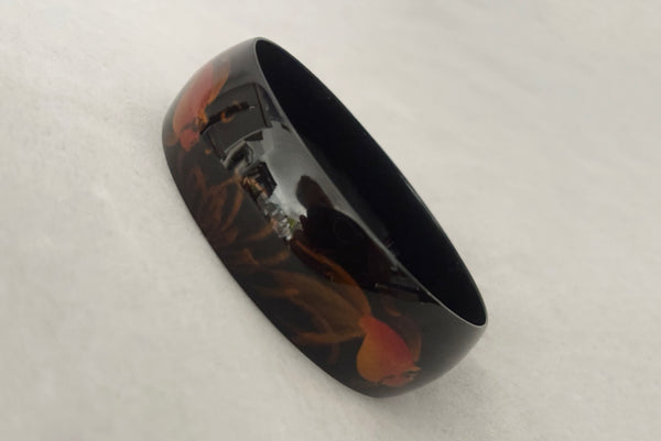 Horizontal perspective view of 1980s vintage black plastic twisted, ridged, and orange goldfish painted bangle bracelet set of three (3). Showing the goldfish painted bangle only.