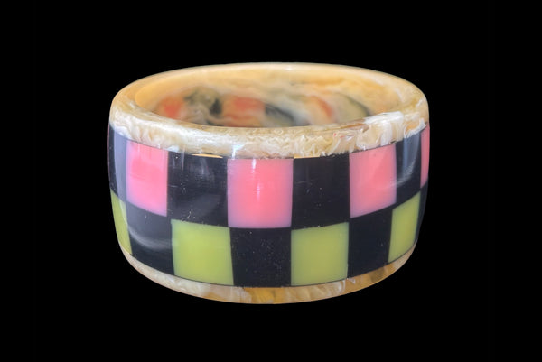 Horizontal front view of 1960s vintage artisan lime green, pink, and black inlaid checkerboard Lucite bangle bracelet on a translucent marbled beige base.