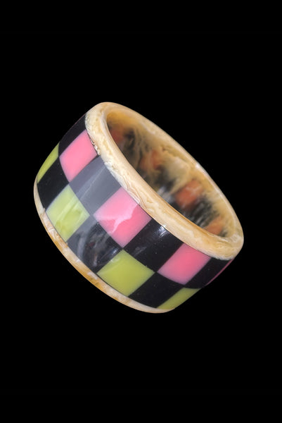 Vertical perspective view of 1960s vintage artisan lime green, pink, and black inlaid checkerboard Lucite bangle bracelet on a translucent marbled beige base.