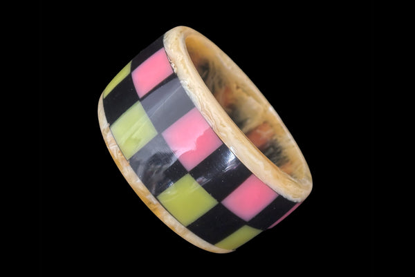 Horizontal perspective view of 1960s vintage artisan lime green, pink, and black inlaid checkerboard Lucite bangle bracelet on a translucent marbled beige base.