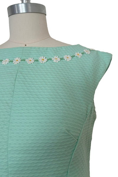 Detail view of 1960s vintage mint green cotton pique daisy trimmed sheath dress, size extra small to small. Showing the boat neckline, daisy trim, and bust darts.