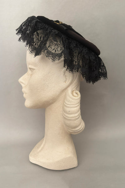 Vertical side view of 1950s vintage black velvet and lace one size pancake cocktail hat with ribbon and gold tone metal buckle, by Lemington for Frederick & Nelson.