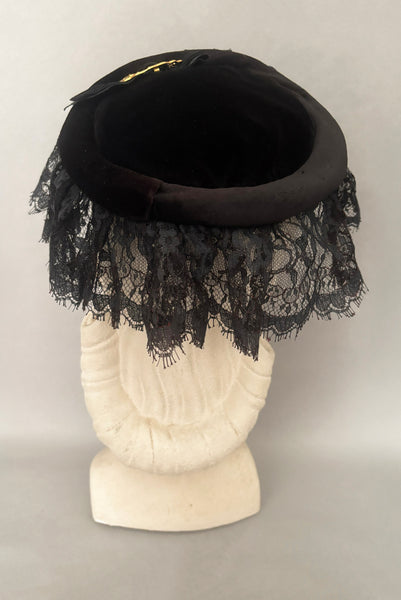 Vertical back view of 1950s vintage black velvet and lace one size pancake cocktail hat with ribbon and gold tone metal buckle, by Lemington for Frederick & Nelson.