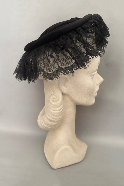 Vertical side view of 1950s vintage black velvet and lace one size pancake cocktail hat with ribbon and gold tone metal buckle, by Lemington for Frederick & Nelson.