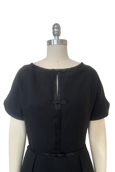 Bodice front view of 1950s vintage black imported linen satin and bow trimmed sheath dress, size small to medium. Showing the round neckline, cap sleeves, and split bodice with satin trim and bows.