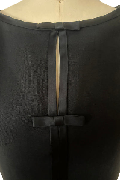 Detail view of 1950s vintage black imported linen satin and bow trimmed sheath dress, size small to medium. Showing the round neckline and split bodice with satin trim and bows.