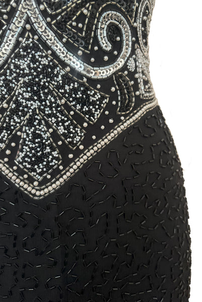 Detail view of 1980s vintage Art Deco faux pearl, silver and black glass beaded black silk chiffon evening dress, size small to medium. With a scoop neckline, sleeveless bodice, and floor length pencil skirt. Showing a close up of the bodice with the silver, white, and black bead and sequin embellished Deco style motif, pointed waistline, and beaded skirt. 