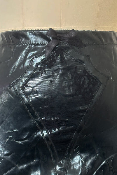 Detail view of 2000s vintage goth pin-up black wet look nylon Lycra stretch lingerie garter skirt by Bernie Dexter, size medium to large. Showing the black elastic at the top edge, black lace embellishment, and black satin bow. 