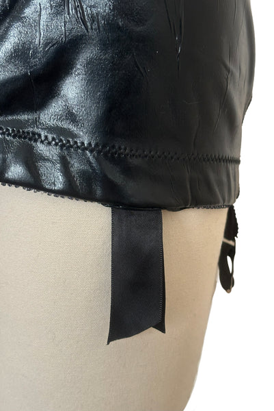 Detail view of 2000s vintage goth pin-up black wet look nylon Lycra stretch lingerie garter skirt by Bernie Dexter, size medium to large. Showing a close-up of the elastic at the bottom edge and the satin ribbon covering the garter clip.