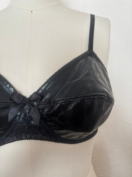 Detail view of 2000s vintage y2k goth pin-up lingerie black nylon Lycra wet look bra by Bernie Dexter for Lucy B, size medium to large. Showing a close-up of the multi-part cup, lace overlay on the stand, and black ribbon bow.