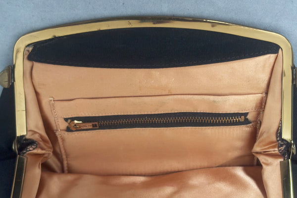Interior detail view of 1950s vintage black faille gold tone metal frame side clasps clutch handbag. Showing the zippered compartment, faded stamp, and discoloration to the peach satin lining.