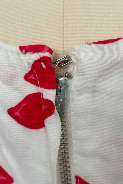 Detail of 1950s vintage red and pink lips novelty border print white cotton sleeveless, full gathered skirt dress, size extra small to small. Showing a close-up of the Green metal zipper.