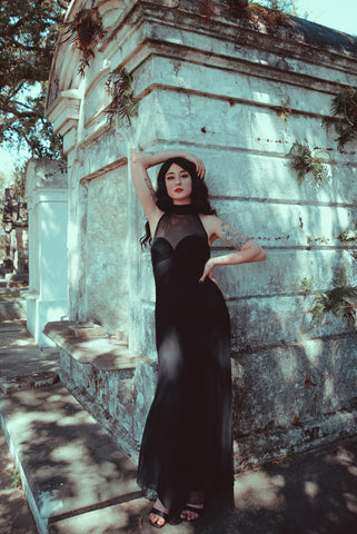 Portrait view of 1990s vintage Tadashi mockneck black mesh maxi length evening dress, small to medium. Showing a model wearing it in a cemetery.